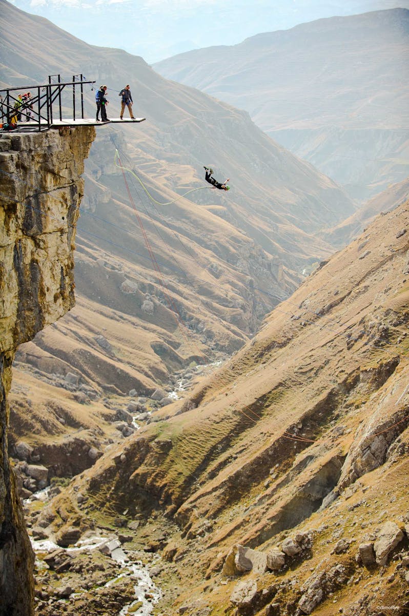 A daring bungee jump over a vast mountainous valley with spectators watching from the cliff edge.
